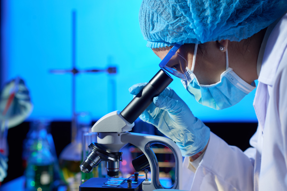 Cientista com máscara e touca olhando através de um microscópio em laboratório.