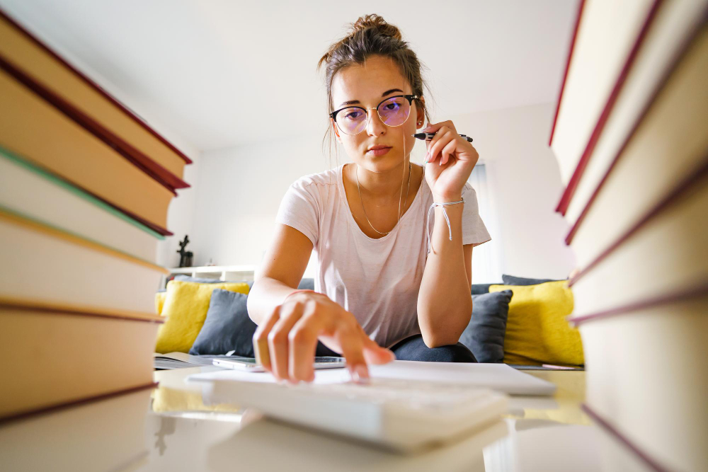 Jovem usando óculos segura caneta e utiliza calculadora, sentada entre pilhas de livros em uma sala de estar com sofá ao fundo.