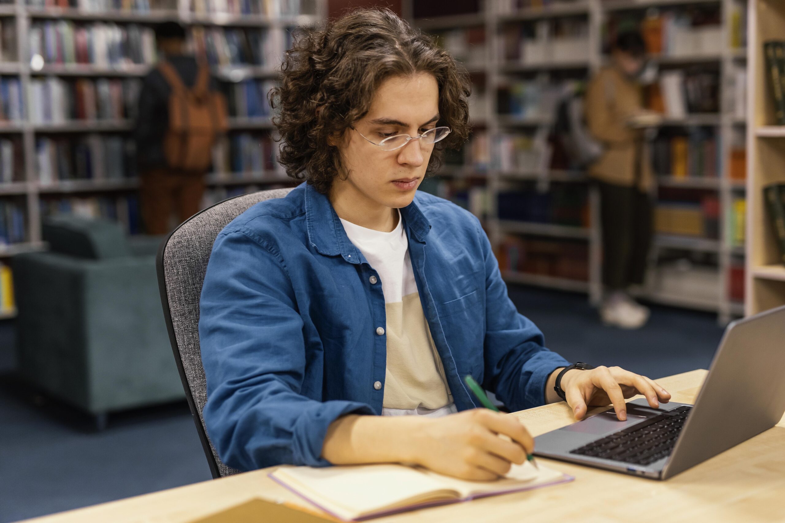Jovem concentrado estudando em uma biblioteca, utilizando um notebook e anotando em um caderno.