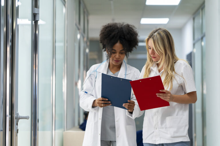 Duas profissionais da saúde, usando jalecos brancos, analisam pranchetas com atenção em um corredor de clínica ou hospital.