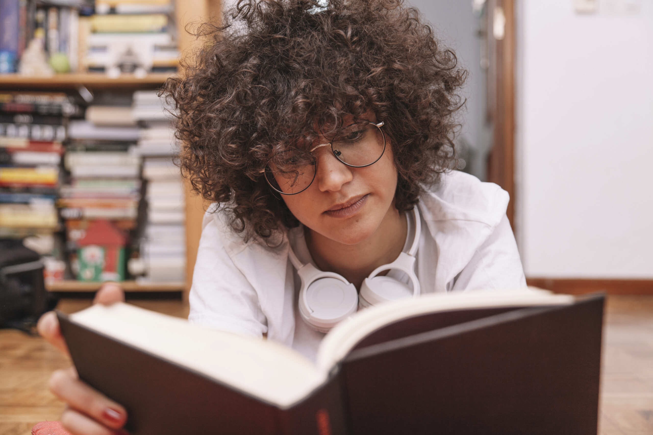 Pessoa de óculos lendo um livro de capa preta no chão, com fones de ouvido no pescoço.