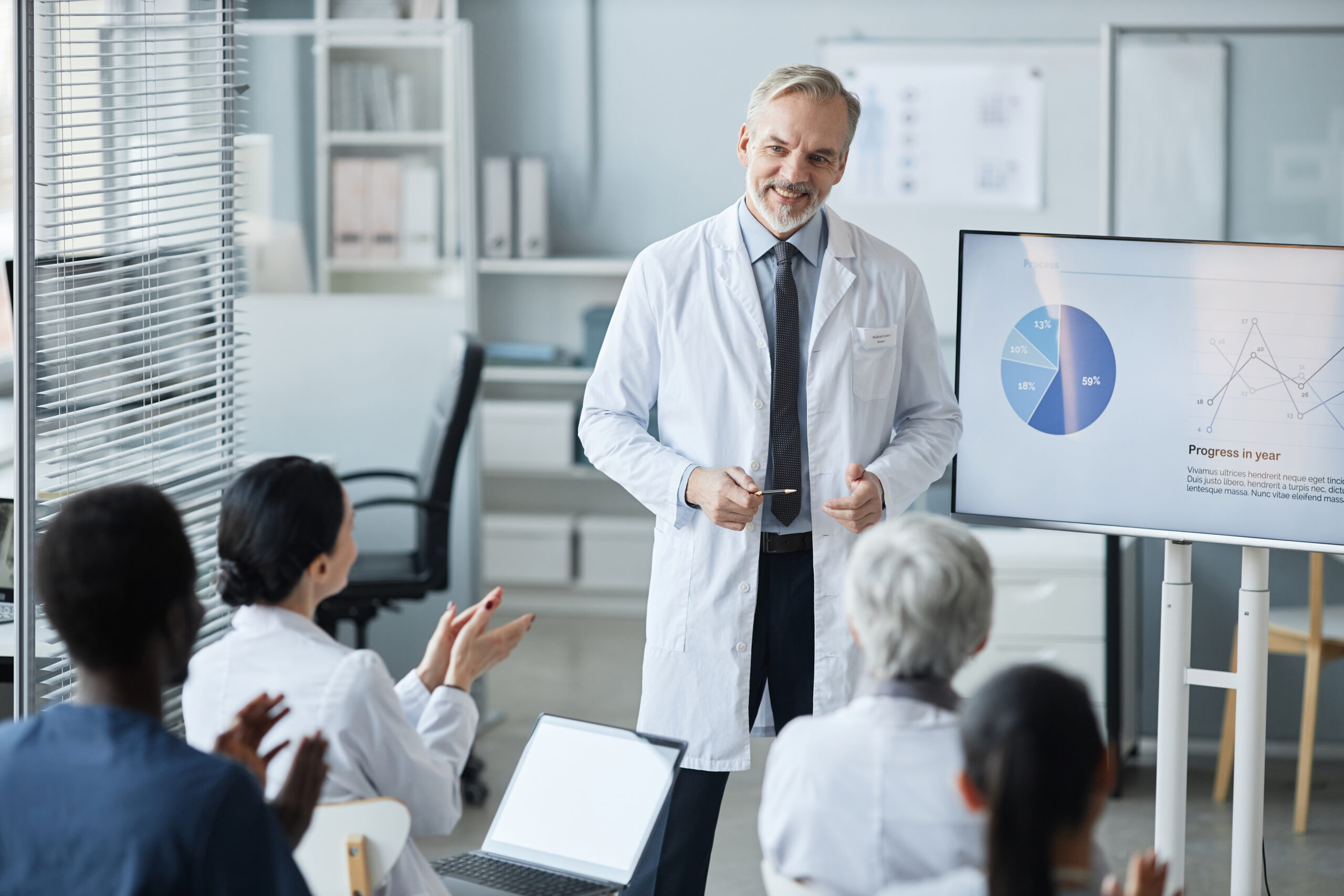 Médico fazendo apresentação com gráficos em uma tela para uma equipe de profissionais da saúde, que o aplaude.