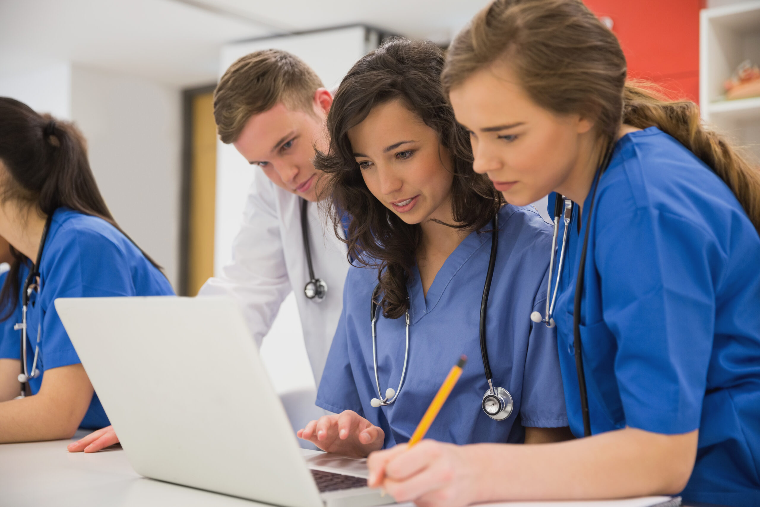 Grupo de estudantes de medicina com jalecos e estetoscópios, reunidos em volta de um notebook, concentrados em um trabalho colaborativo.