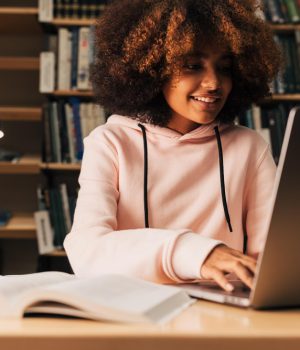 Uma jovem estudante, usando moletom rosa, está sentada em uma biblioteca estudando com o notebook aberto.