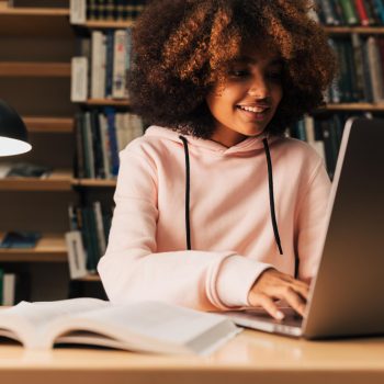 Uma jovem estudante, usando moletom rosa, está sentada em uma biblioteca estudando com o notebook aberto.
