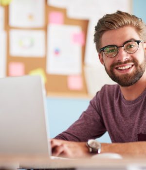Homem jovem sorrindo enquanto trabalha em um laptop, com quadro de anotações ao fundo.