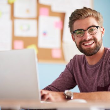 Homem jovem sorrindo enquanto trabalha em um laptop, com quadro de anotações ao fundo.