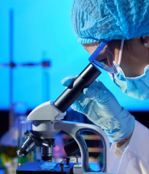 Cientista com máscara e touca olhando através de um microscópio em laboratório.