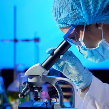 Cientista com máscara e touca olhando através de um microscópio em laboratório.