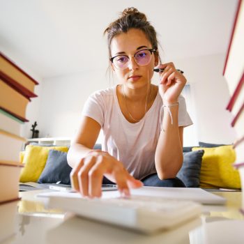 Jovem usando óculos segura caneta e utiliza calculadora, sentada entre pilhas de livros em uma sala de estar com sofá ao fundo.