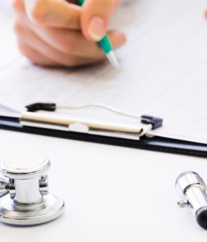Estetoscópio e martelo neurológico sobre mesa médica enquanto profissional preenche formulário clínico em prancheta.