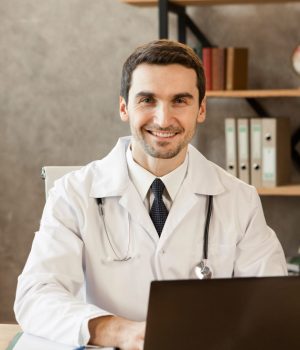 Profissional de saúde sorridente, usando jaleco branco e estetoscópio, sentado à mesa com notebook em consultório.