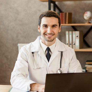 Profissional de saúde sorridente, usando jaleco branco e estetoscópio, sentado à mesa com notebook em consultório.