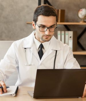 Médico usando jaleco e óculos trabalha em um consultório, olhando para um notebook e anotando em um caderno.