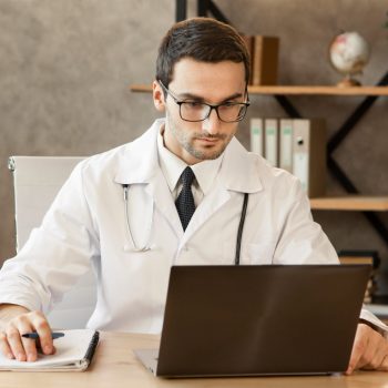 Médico usando jaleco e óculos trabalha em um consultório, olhando para um notebook e anotando em um caderno.