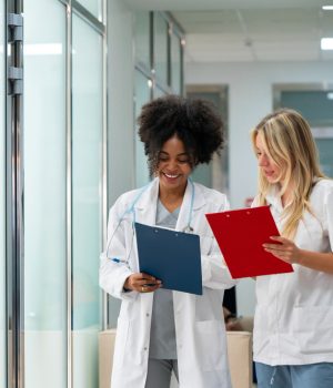 Duas profissionais da saúde conversando em corredor de clínica ou hospital enquanto analisam pranchetas médicas.