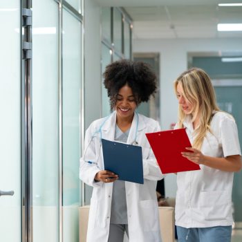 Duas profissionais da saúde conversando em corredor de clínica ou hospital enquanto analisam pranchetas médicas.