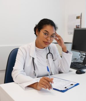 Uma médica jovem, de jaleco branco e estetoscópio no pescoço, está sentada à mesa de um consultório, segurando uma caneta e olhando para a câmera.