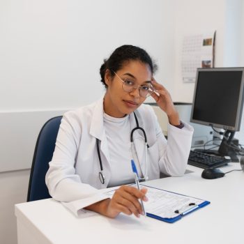 Uma médica jovem, de jaleco branco e estetoscópio no pescoço, está sentada à mesa de um consultório, segurando uma caneta e olhando para a câmera.