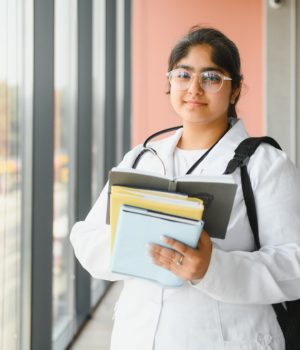 Estudante de Medicina com jaleco branco e livros nas mãos, em um corredor iluminado por janelas grandes, simbolizando o aprendizado e a rotina médica.
