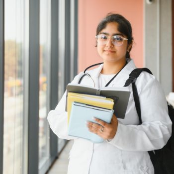 Estudante de Medicina com jaleco branco e livros nas mãos, em um corredor iluminado por janelas grandes, simbolizando o aprendizado e a rotina médica.