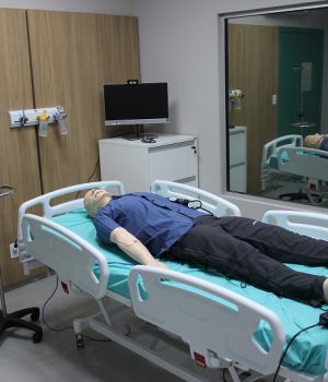 Boneco anatômico de simulação médica deitado em uma cama hospitalar dentro de uma sala equipada.