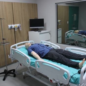 Boneco anatômico de simulação médica deitado em uma cama hospitalar dentro de uma sala equipada.