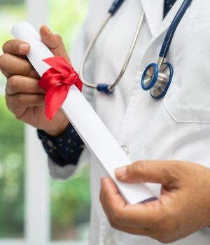Profissional da saúde usando jaleco e estetoscópio segurando diploma enrolado com laço vermelho.