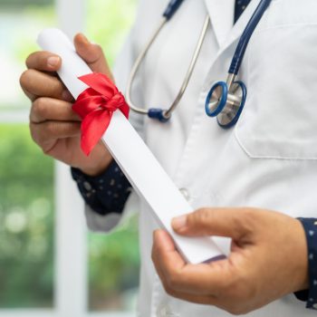 Profissional da saúde usando jaleco e estetoscópio segurando diploma enrolado com laço vermelho.