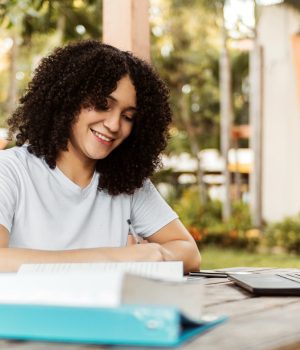 Jovem sorridente estuda ao ar livre, escrevendo em um caderno sobre a mesa com livros e laptop abertos.