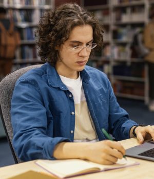 Jovem concentrado estudando em uma biblioteca, utilizando um notebook e anotando em um caderno.
