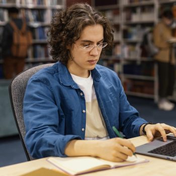 Jovem concentrado estudando em uma biblioteca, utilizando um notebook e anotando em um caderno.