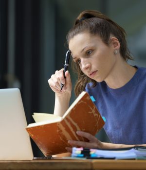 Jovem mulher estudando com um caderno e um notebook, concentrada e pensativa.