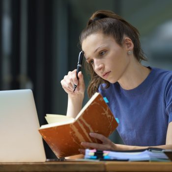 Jovem mulher estudando com um caderno e um notebook, concentrada e pensativa.
