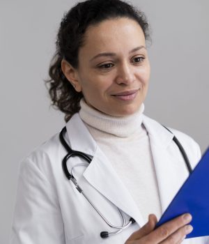 Médica com jaleco e estetoscópio olha atentamente para uma prancheta azul, em ambiente iluminado por luz natural.