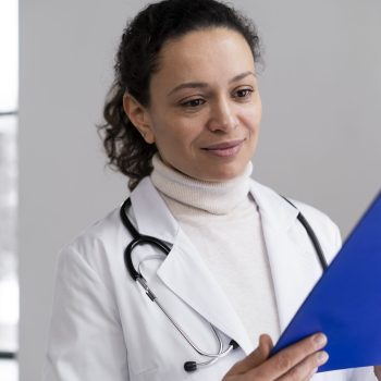 Médica com jaleco e estetoscópio olha atentamente para uma prancheta azul, em ambiente iluminado por luz natural.