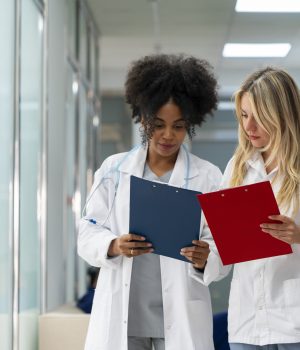 Duas profissionais da saúde, usando jalecos brancos, analisam pranchetas com atenção em um corredor de clínica ou hospital.