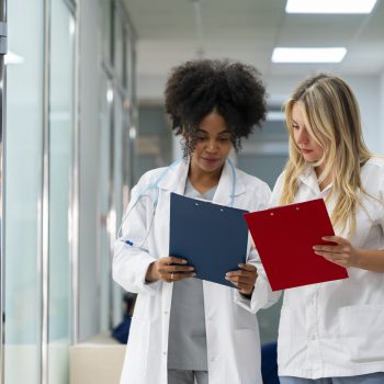 Duas profissionais da saúde, usando jalecos brancos, analisam pranchetas com atenção em um corredor de clínica ou hospital.