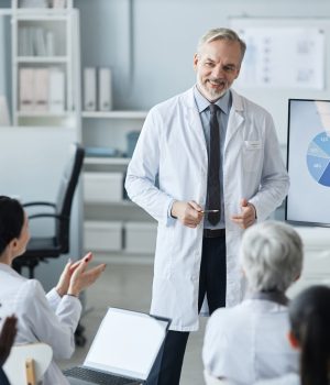 Médico fazendo apresentação com gráficos em uma tela para uma equipe de profissionais da saúde, que o aplaude.
