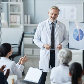 Médico fazendo apresentação com gráficos em uma tela para uma equipe de profissionais da saúde, que o aplaude.