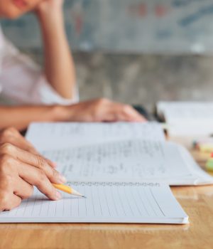 Pessoa escrevendo em um caderno com lápis enquanto estuda, com livros e marcadores de texto sobre a mesa