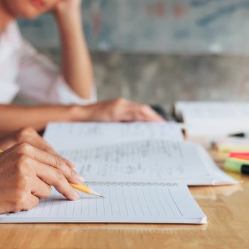 Pessoa escrevendo em um caderno com lápis enquanto estuda, com livros e marcadores de texto sobre a mesa
