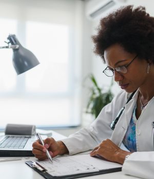 Uma médica usando um jaleco branco e óculos está sentada em uma mesa de escritório, escrevendo em uma prancheta. Há um notebook e uma luminária na mesa.