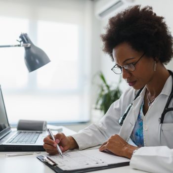 Uma médica usando um jaleco branco e óculos está sentada em uma mesa de escritório, escrevendo em uma prancheta. Há um notebook e uma luminária na mesa.