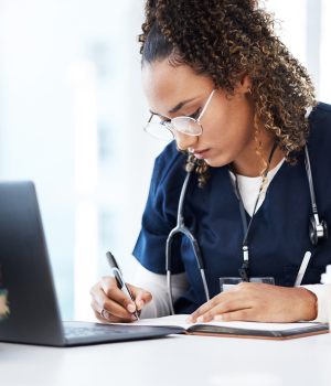 Uma jovem mulher com cabelos cacheados e óculos, vestindo um uniforme médico azul (scrubs) e com um estetoscópio ao redor do pescoço, está sentada em uma mesa. Ela está concentrada, escrevendo em um caderno aberto com uma caneta. Em frente a ela, há um laptop aberto e uma pilha de livros