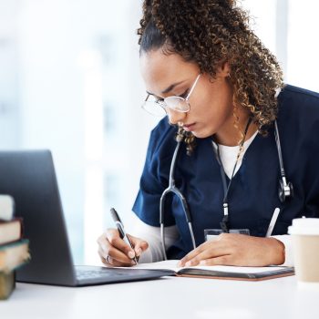 Uma jovem mulher com cabelos cacheados e óculos, vestindo um uniforme médico azul (scrubs) e com um estetoscópio ao redor do pescoço, está sentada em uma mesa. Ela está concentrada, escrevendo em um caderno aberto com uma caneta. Em frente a ela, há um laptop aberto e uma pilha de livros
