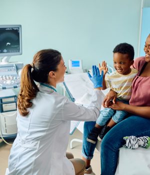 Médica com luvas azuis interagindo com um menino sorridente sentado no colo da mãe em um consultório pediátrico.