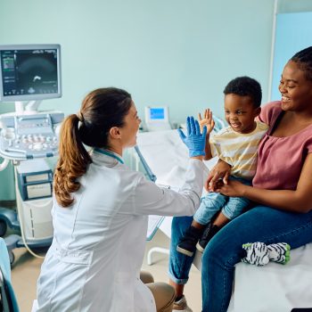 Médica com luvas azuis interagindo com um menino sorridente sentado no colo da mãe em um consultório pediátrico.