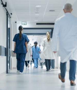 Profissionais de saúde caminham por corredor de hospital moderno, usando jalecos e uniformes clínicos.