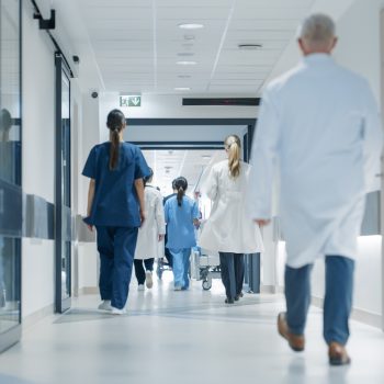 Profissionais de saúde caminham por corredor de hospital moderno, usando jalecos e uniformes clínicos.
